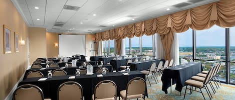 Un salón de conferencias con varias mesas y sillas, ventanas amplias con cortinas y vistas a la ciudad.
