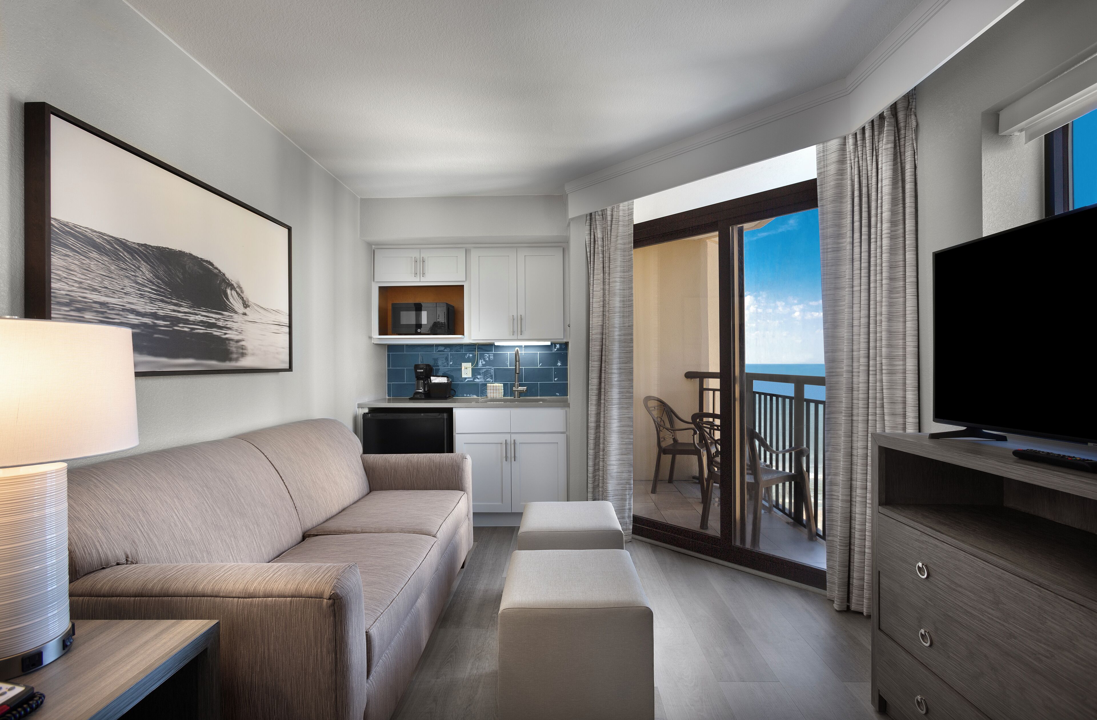 Una habitación de hotel moderna con sofá, televisor, balcón con vistas y zona de cocina.