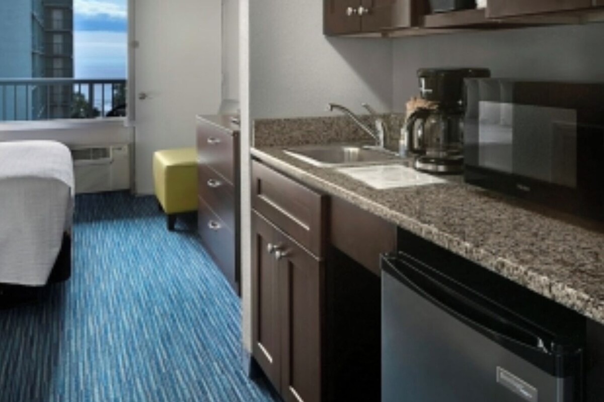 Une chambre d’hôtel avec un lit, une petite kitchenette, une fenêtre donnant sur des immeubles et un balcon.