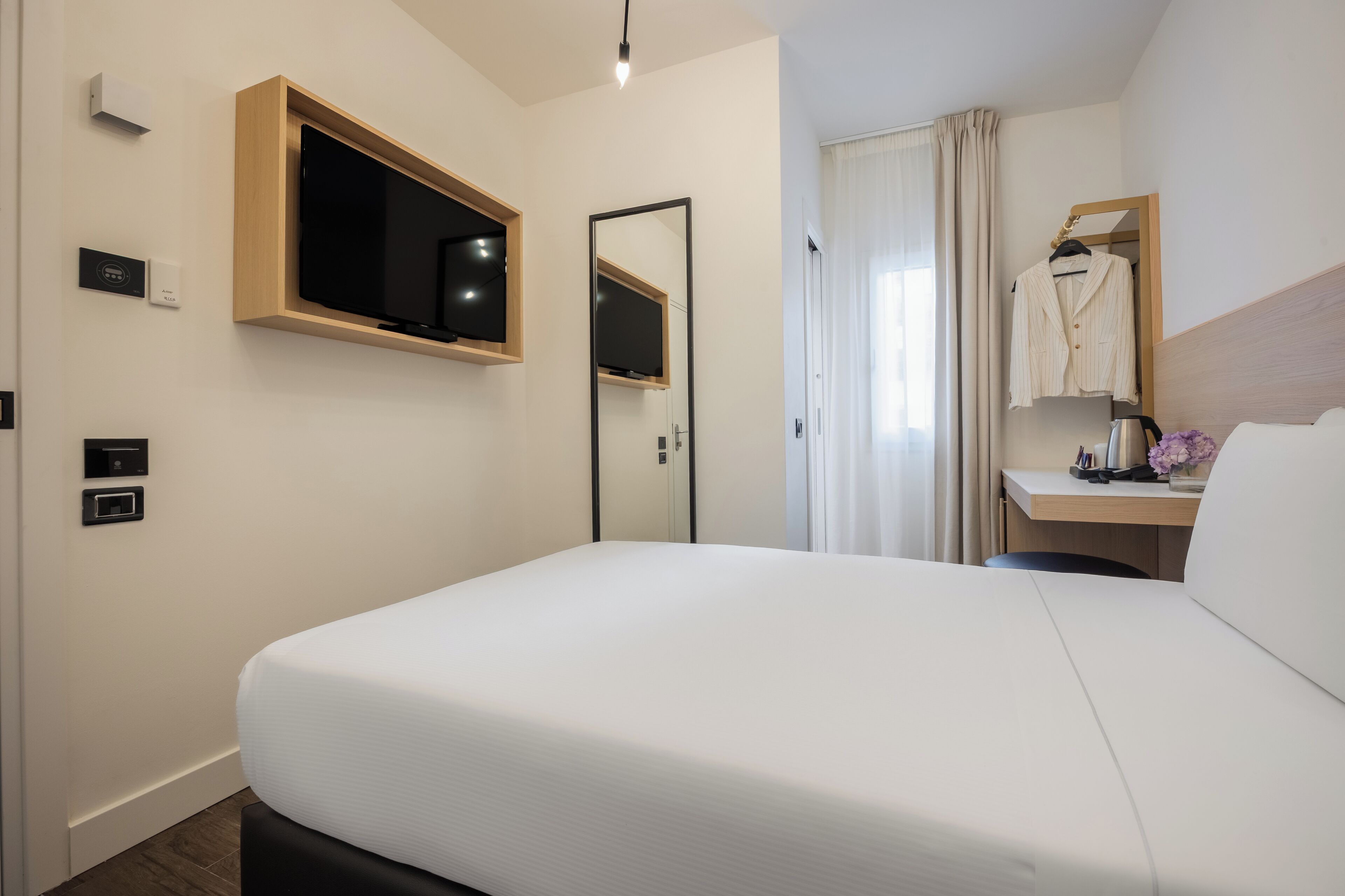Een hotelkamer met een groot bed, een aan de muur bevestigde televisie, een spiegel, een bureau met stoel, en een raam met gordijnen.