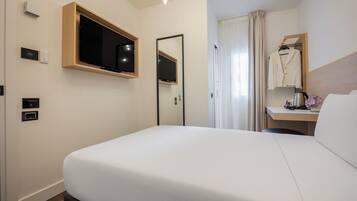 Een hotelkamer met een groot bed, een aan de muur bevestigde televisie, een spiegel, een bureau met stoel, en een raam met gordijnen.