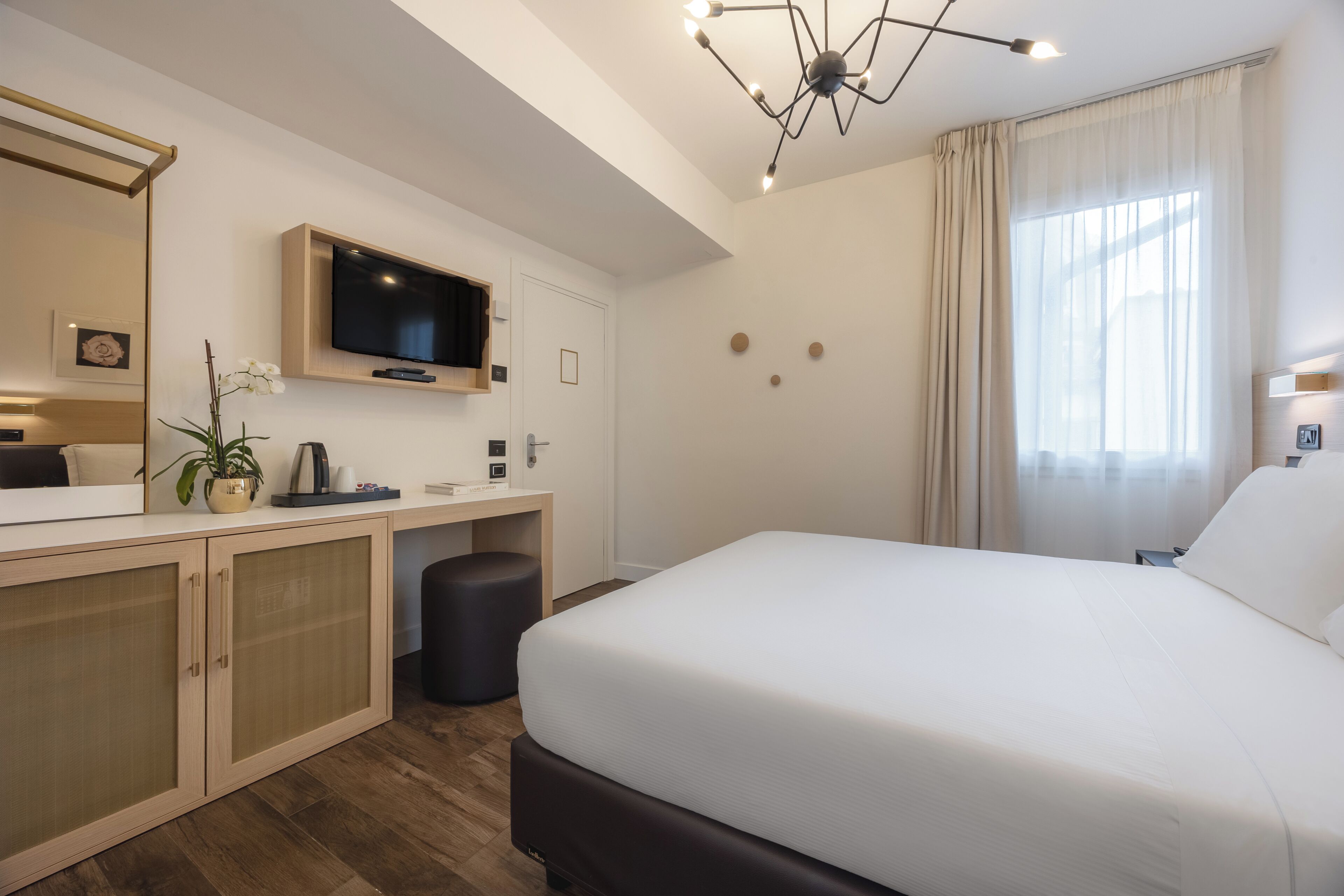 Camera d'albergo moderna con un ampio letto, una scrivania con televisore, uno specchio e una pianta in vaso.