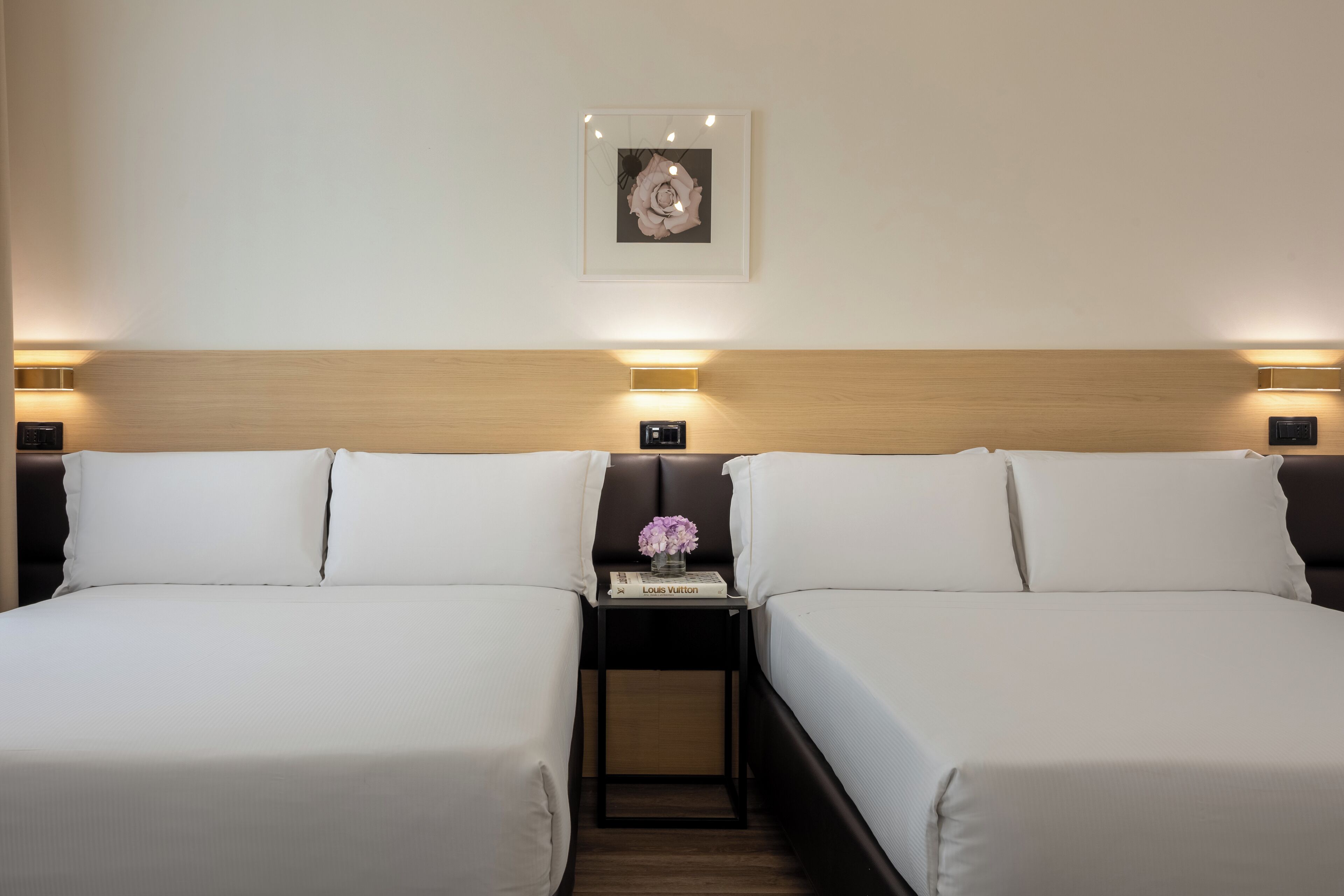 A hotel room with two beds, a nightstand with a vase of flowers, a framed picture on the wall, and wall-mounted lights.