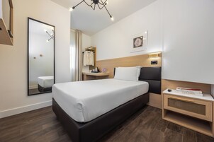 Una habitación de hotel moderna con una cama grande, una mesita de noche con lámpara, un espejo, un pequeño aparador y una mesita.