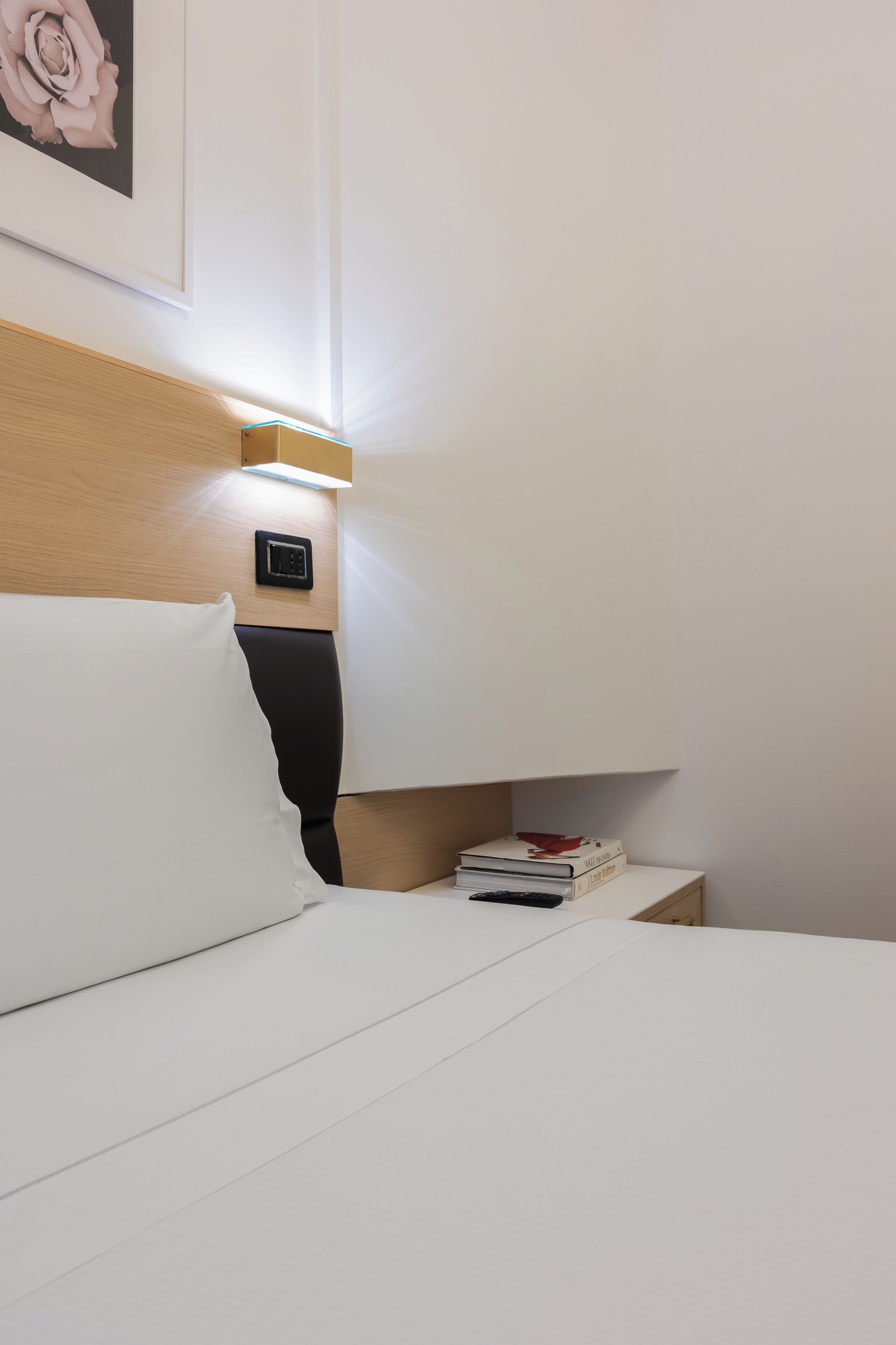 A neatly made bed with white linens, a bedside table with a stack of books, a wall-mounted light, and a framed picture of a rose.