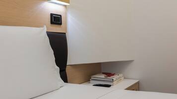 Una cama bien hecha con sábanas blancas, una mesita de noche con una pila de libros, una lámpara de pared y un cuadro con una rosa.