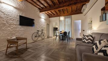Amplia sala de estar con piso de madera en patrón de espiga, un sofá con cojines estampados, una silla de mimbre, una mesa de comedor con sillas y una bicicleta apoyada en la pared.