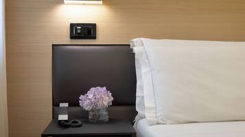 Quarto de hotel bem arrumado com uma cama, mesa de cabeceira com um vaso de flores, luminária e painel de controle na parede.