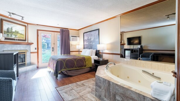 Habitación de hotel con una cama grande, una zona de estar con una silla, bañera, televisor y un ventanal con cortinas.