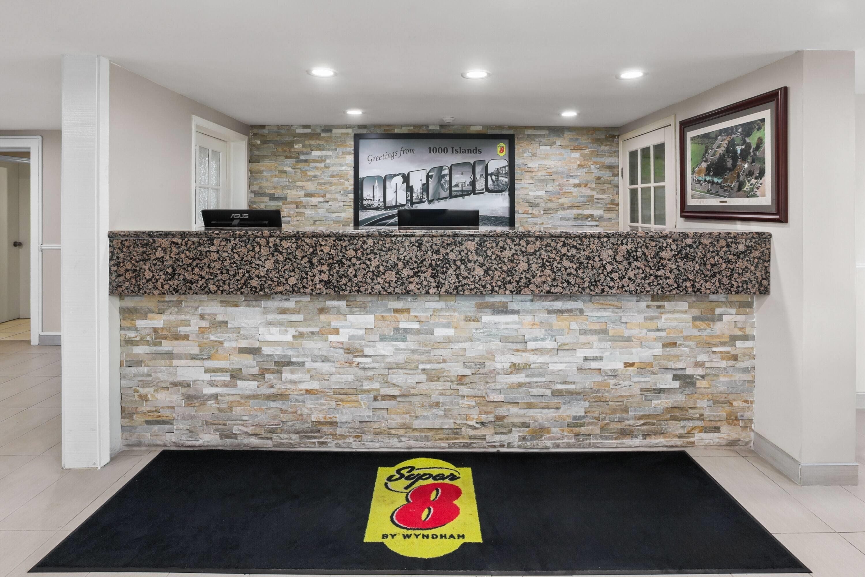 Un área de recepción con una pared de piedra, alfombra negra con un logotipo, un cuadro enmarcado y una computadora portátil sobre el mostrador.