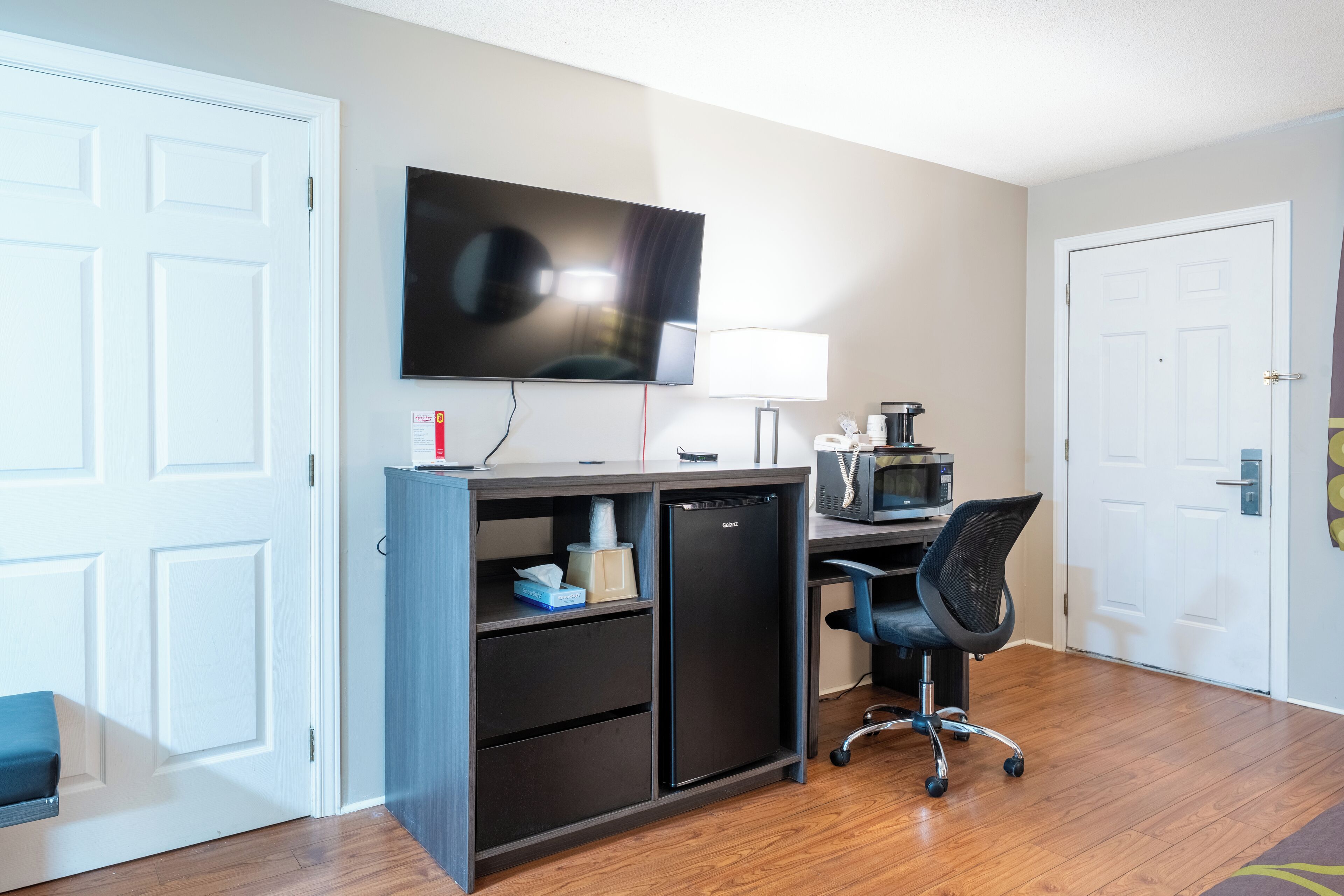 Habitación de hotel con escritorio, televisión de pantalla plana, microondas, refrigerador, silla y puerta.