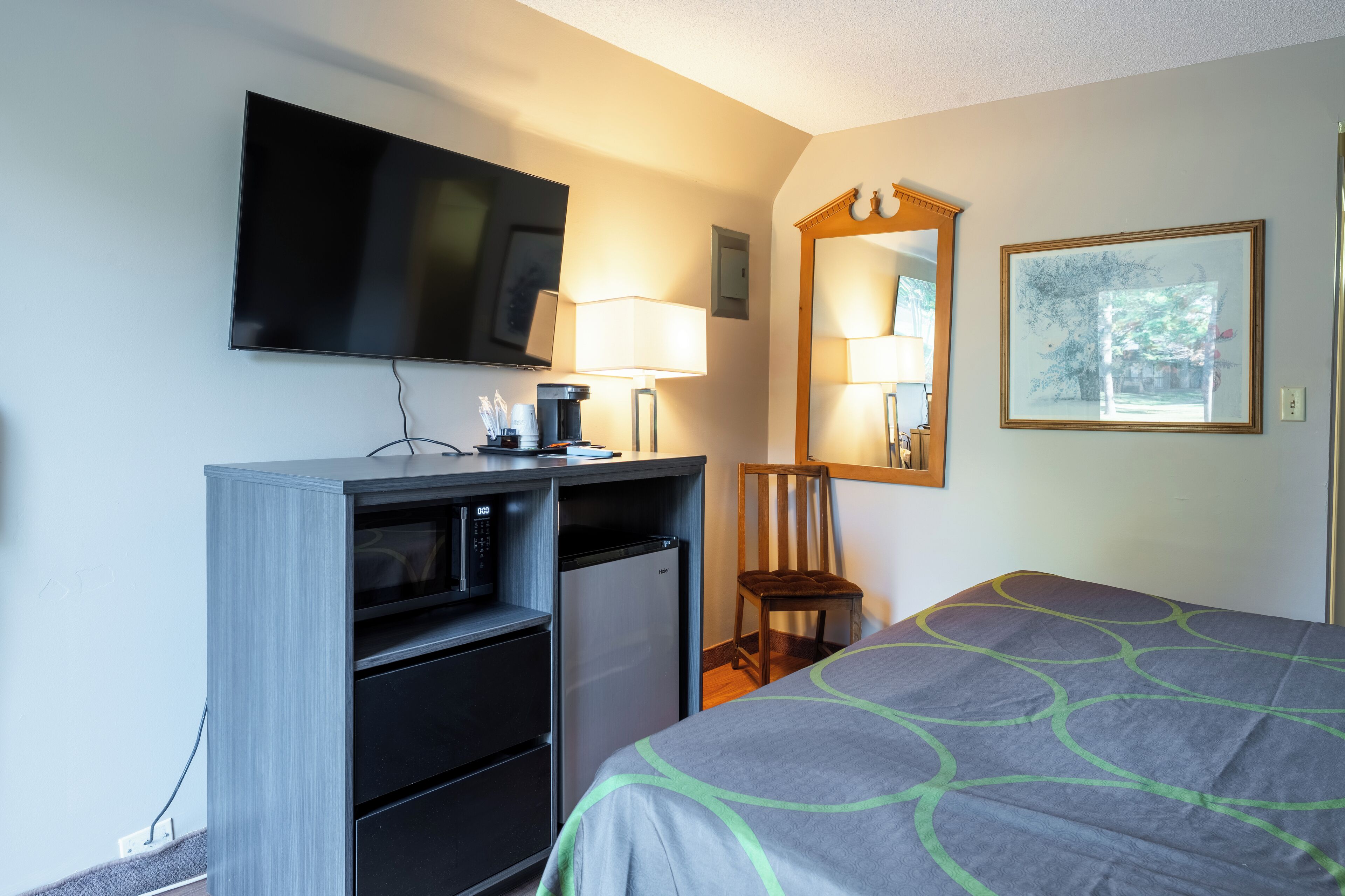 A hotel room with a bed, a desk with a television, a refrigerator, a chair, a lamp, and a framed picture on the wall.