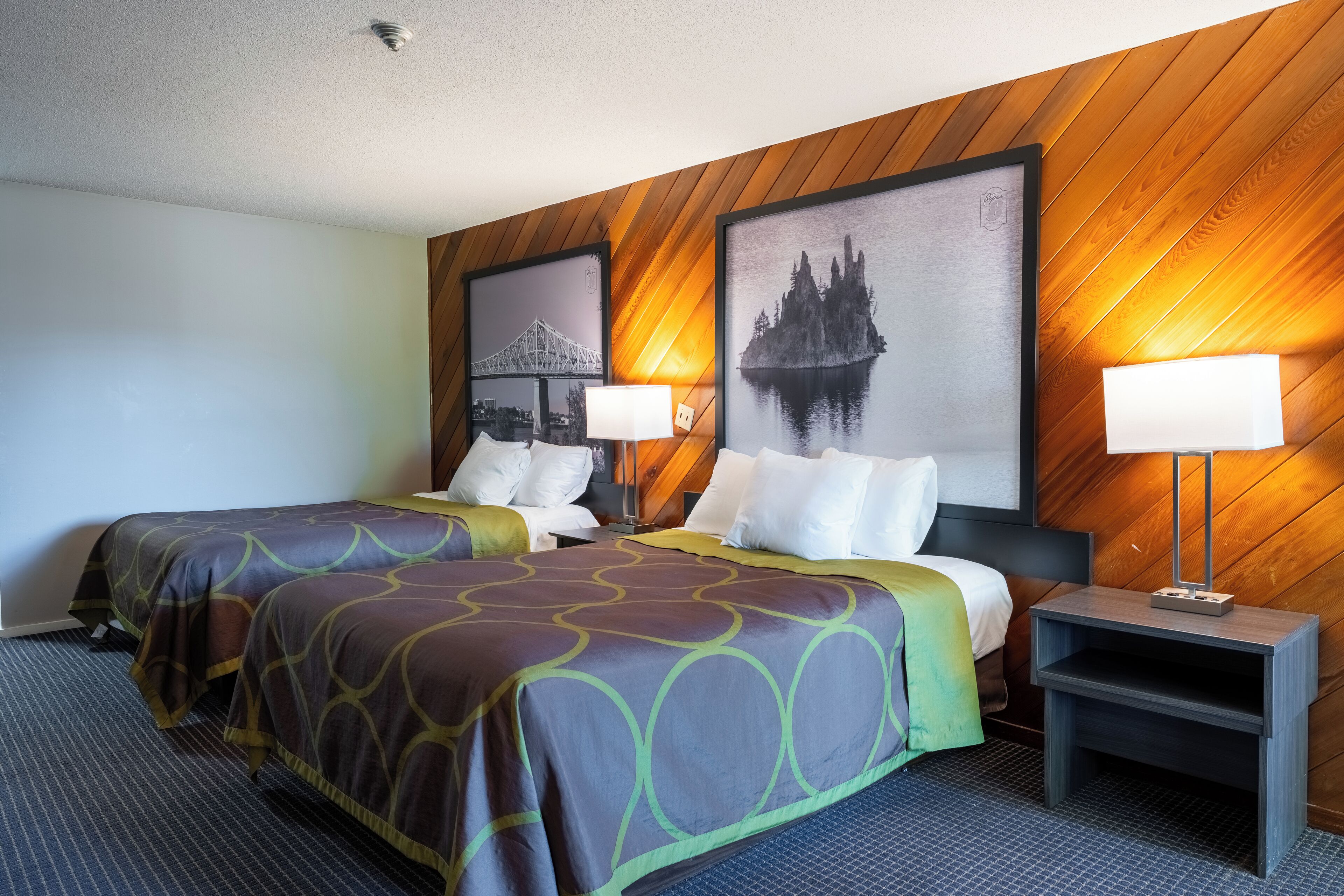 A hotel room with two beds, a nightstand, and a wall-mounted lamp.