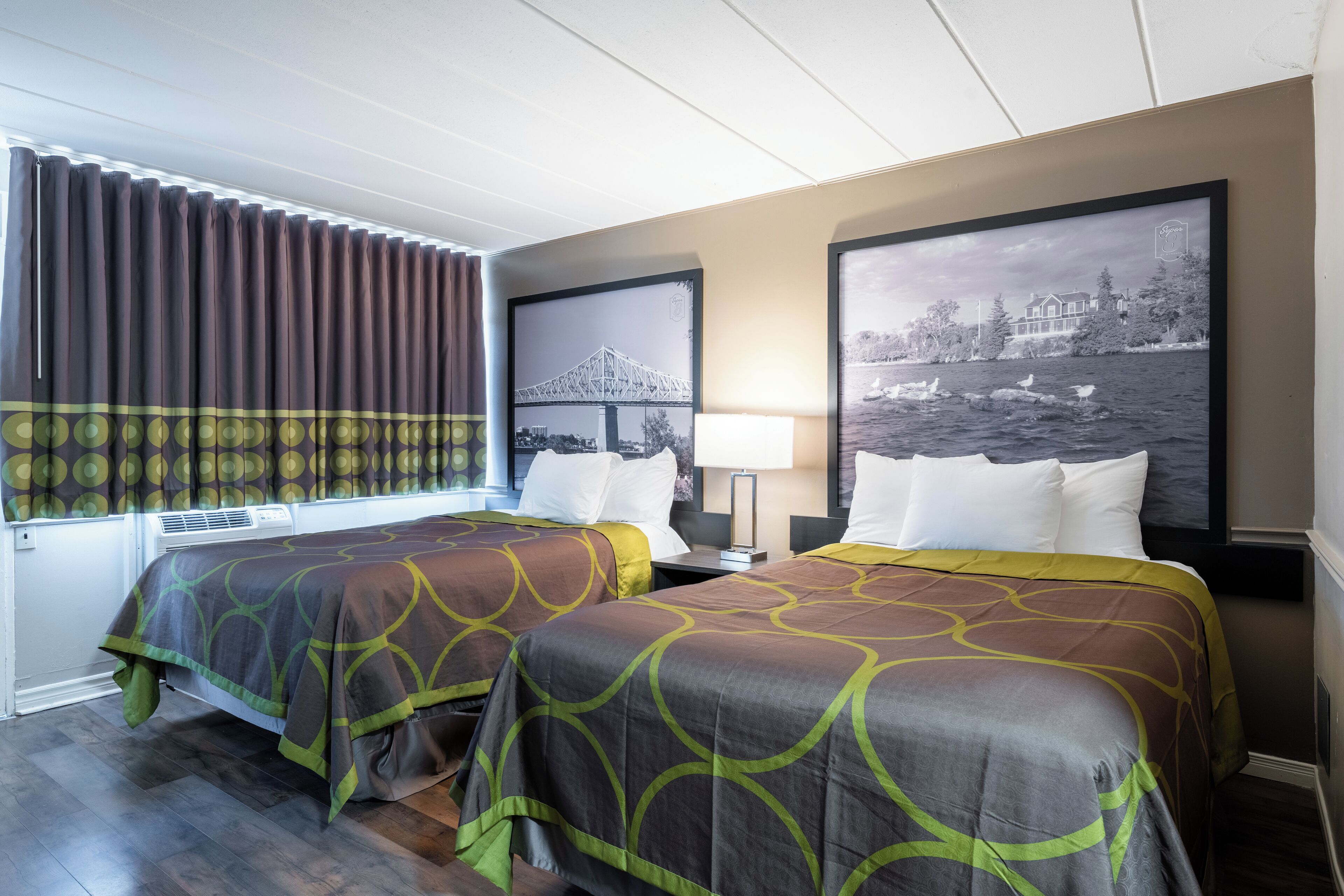 A hotel room with two beds, a window with curtains, a wall-mounted air conditioner, and two framed pictures.