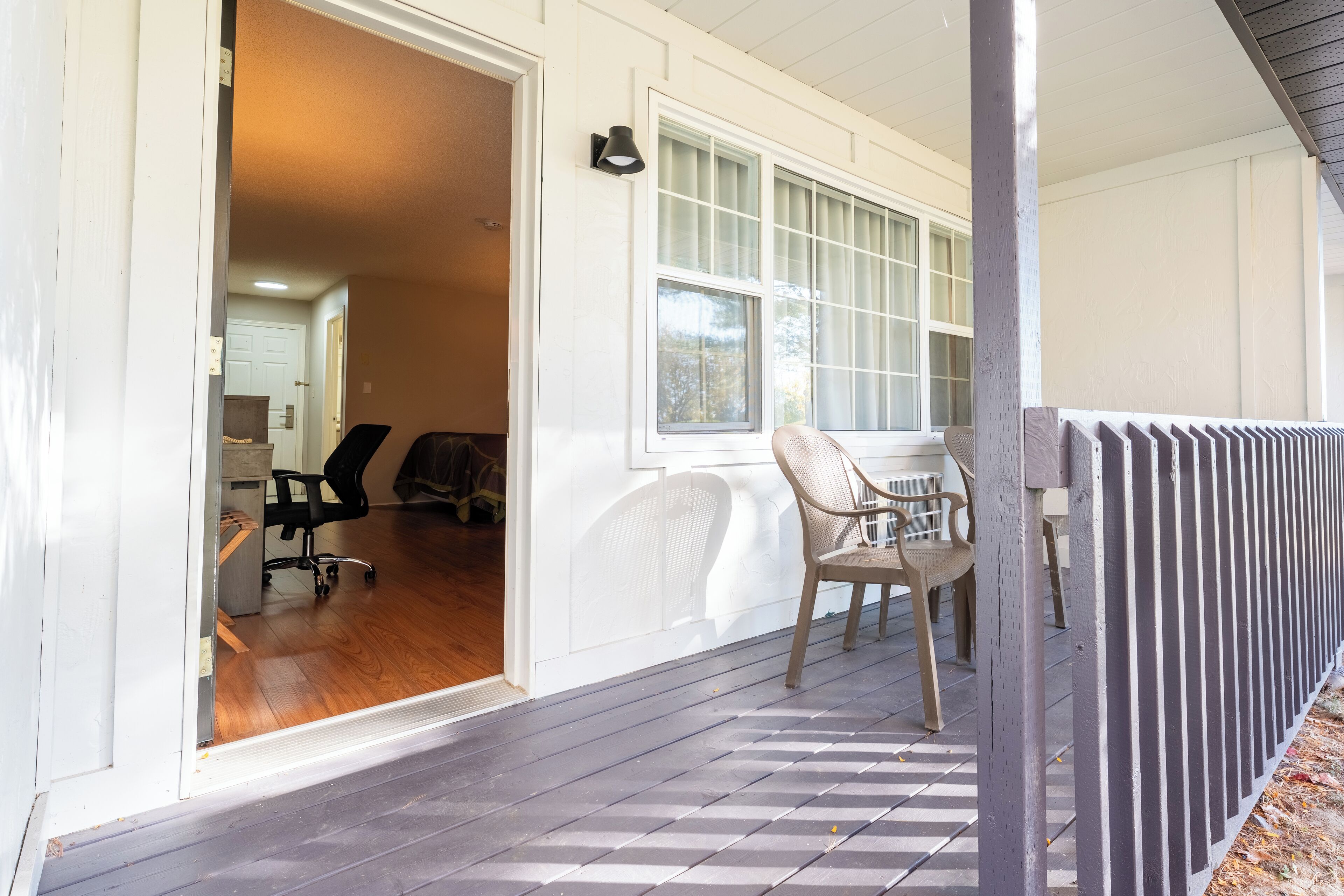 A covered porch with a wooden floor, a white exterior wall, and a window with blinds.