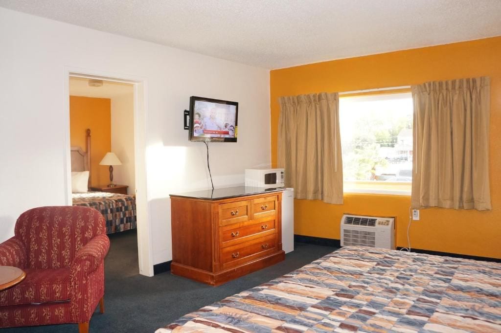 Habitación de hotel con cama, cómoda de madera, televisor montado en la pared, ventana con cortinas y un sillón rojo.
