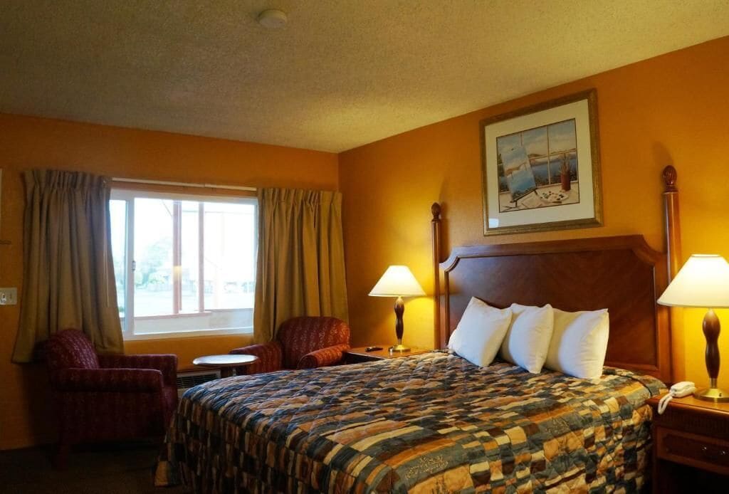 A hotel room with a large bed, two bedside lamps, a chair, a small table, and a window with curtains.