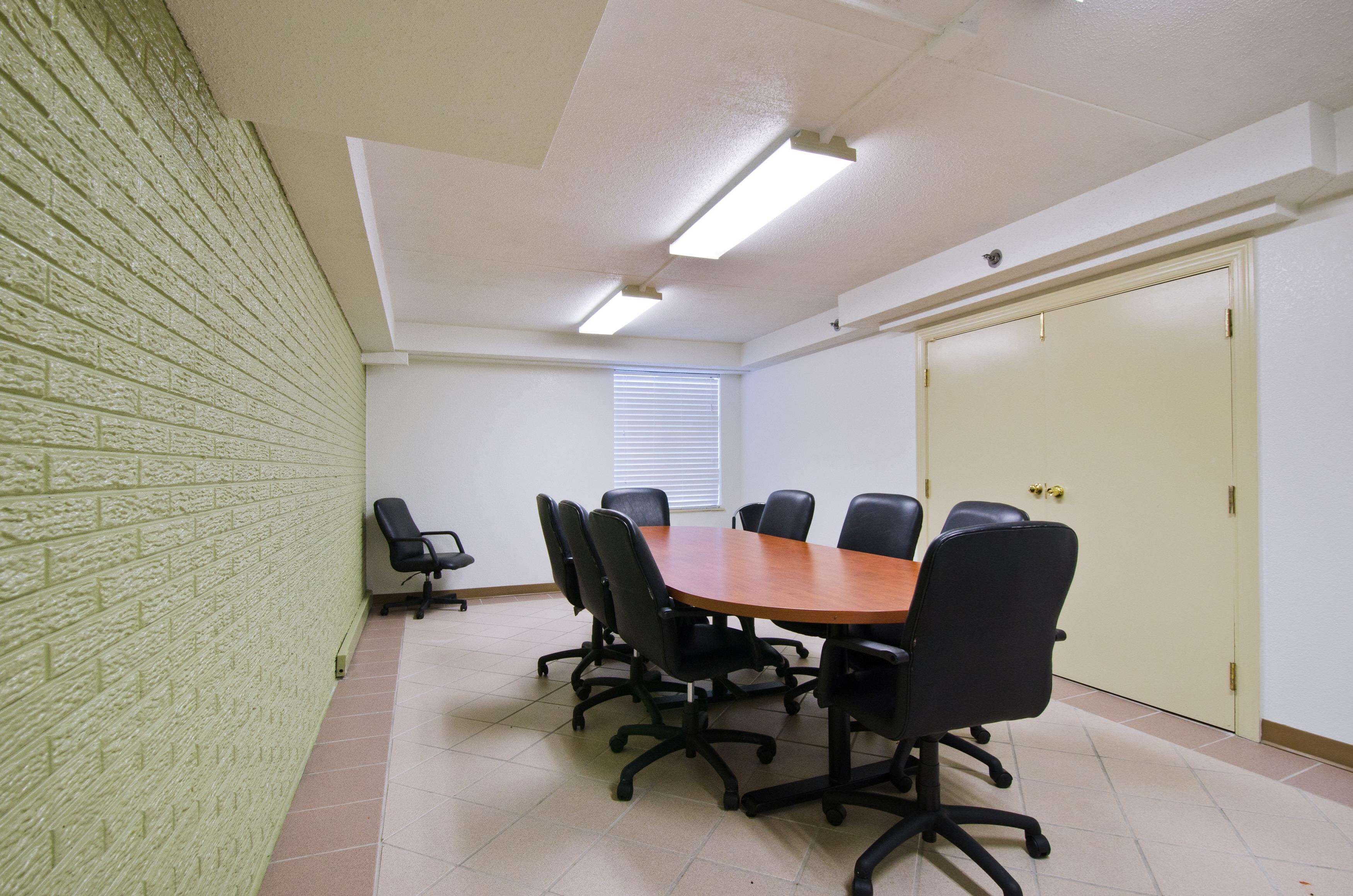 A conference room with a rectangular table, six black chairs, a white ceiling with fluorescent lights, a brick wall, a window with blinds, and a door with a handle.