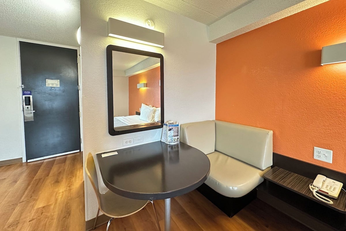 Une chambre d’hôtel avec un plancher en bois, une petite table avec une chaise, un canapé, un miroir et un téléphone posé sur une étagère.