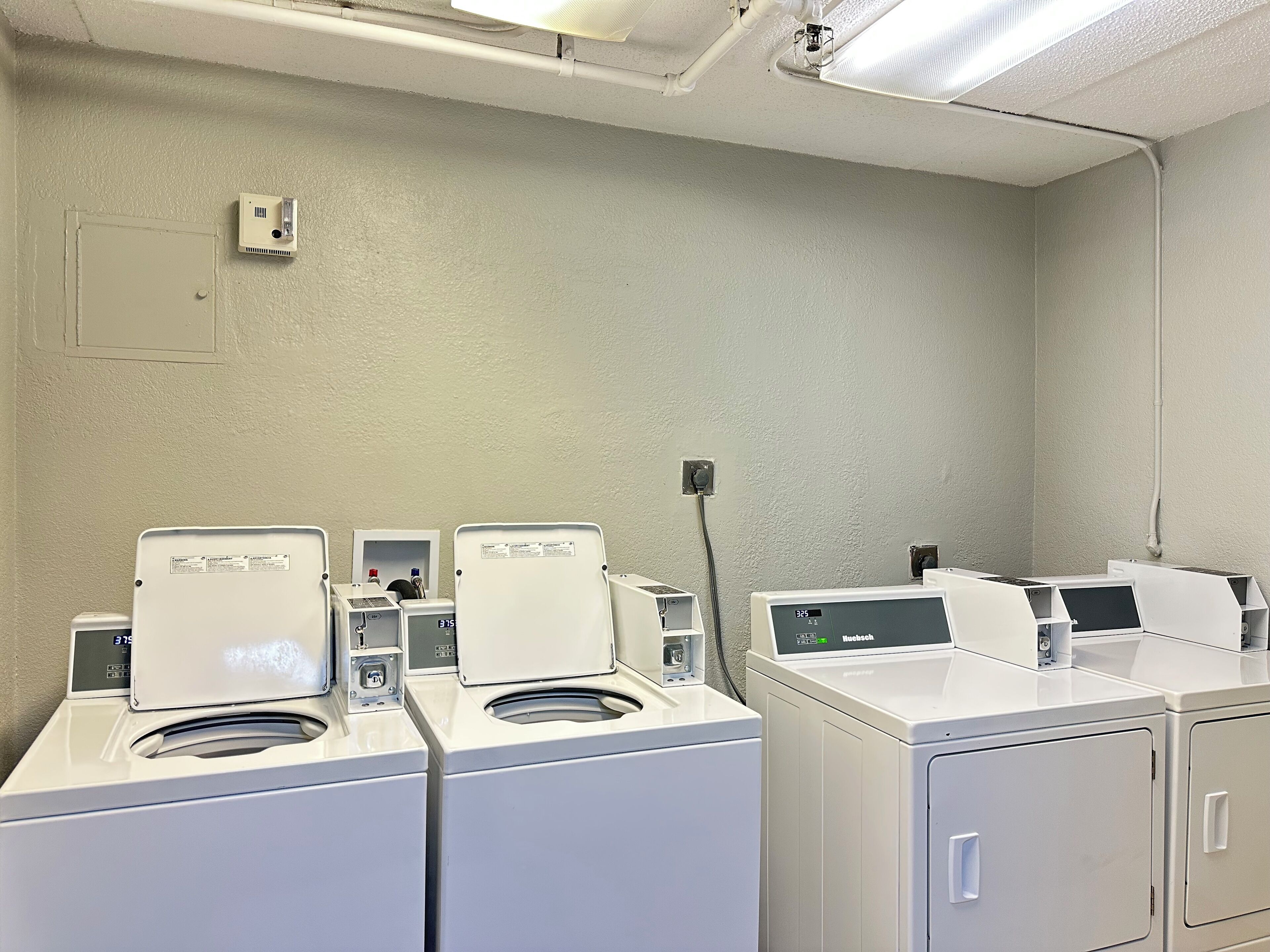 Une buanderie équipée de plusieurs lave-linge et sèche-linge à chargement frontal, d’une prise de courant au mur et d’une bouche d’aération au plafond.