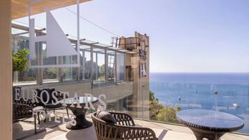 Un balcón con mobiliario de mimbre, una mesa de cristal y vista al mar.