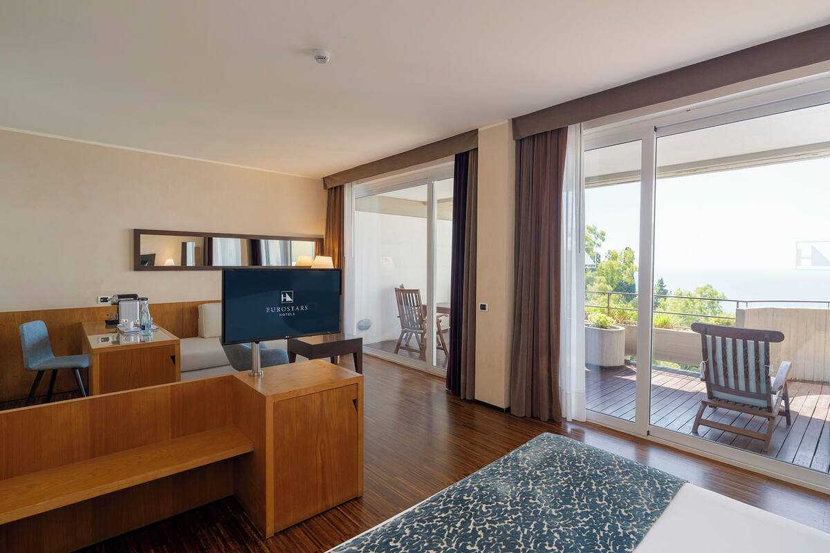 Habitación de hotel con escritorio de madera, televisor, sofá y balcón con mobiliario exterior.
