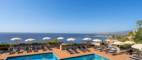 Zona junto a la piscina con sillones y sombrillas, con vista a un mar azul cristalino y tierra distante.