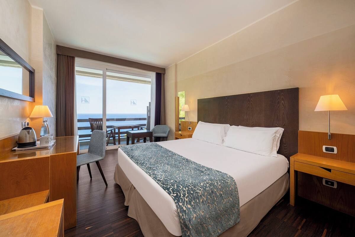 Habitación de hotel con una cama grande, un escritorio con lámpara, una silla y vistas al mar.