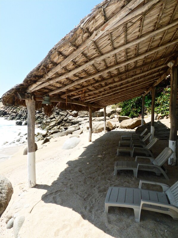 Una estructura junto a la playa con techo de paja y pilares de madera, que incluye varias sillas de descanso.