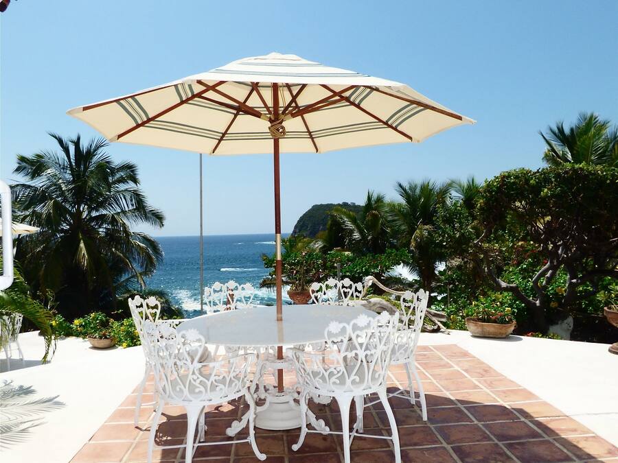 A patio with a round table and white chairs, an umbrella, and a view of the ocean.