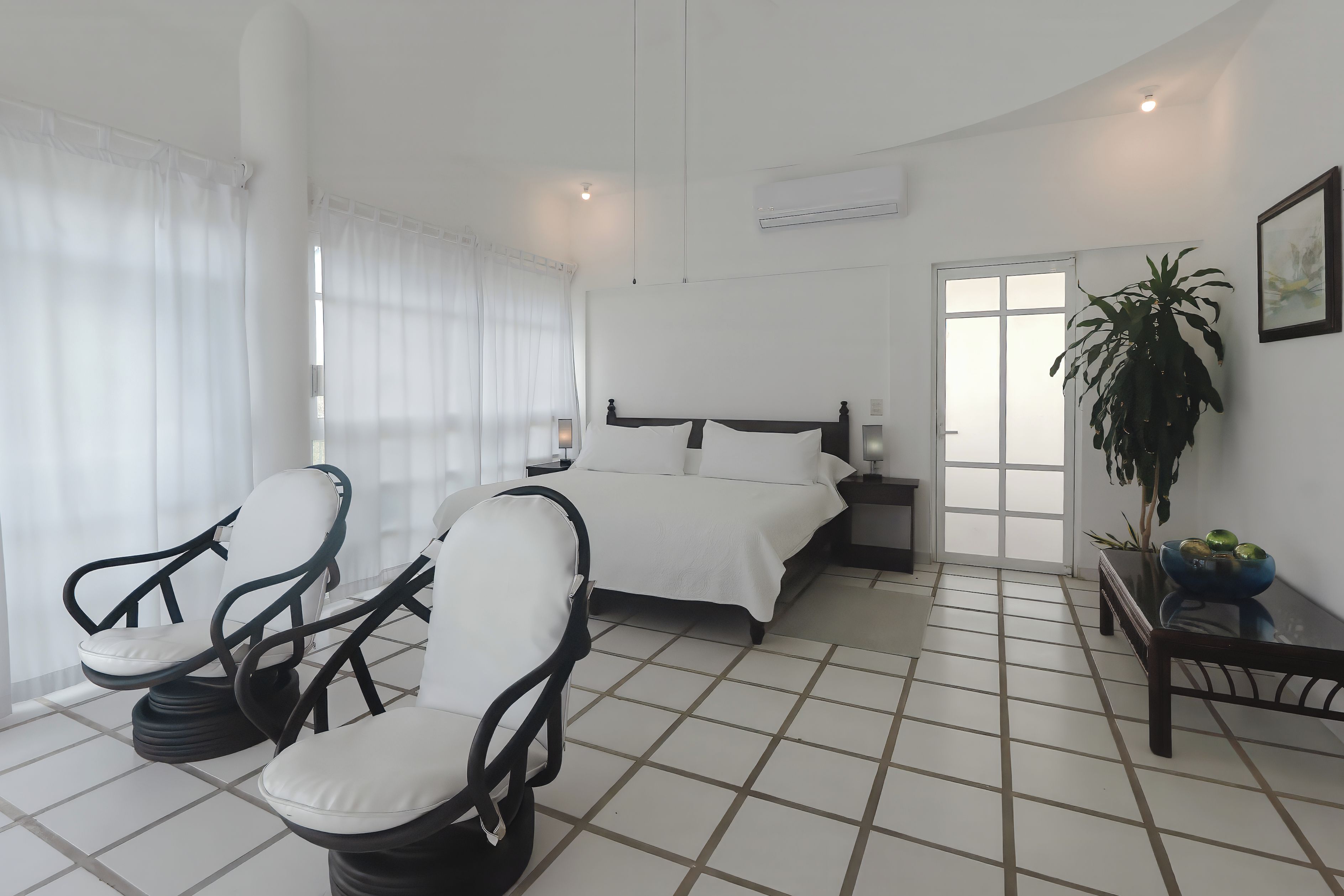 A hotel room with a bed, two chairs, a bedside table, a potted plant, and a window with curtains.