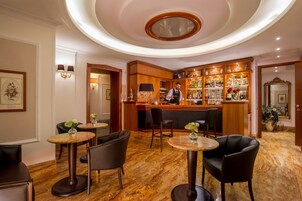 A bar area with a bartender, wooden bar counter, and shelves of bottles. There are round tables with chairs, and a potted plant.