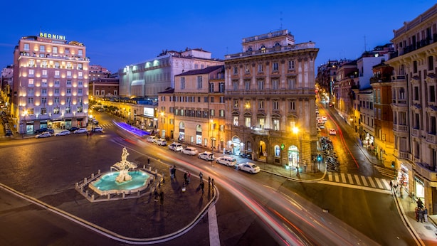 Uma praça histórica com uma fonte central, rodeada por arquitetura europeia clássica e um hotel chamado 'BERNINI'.