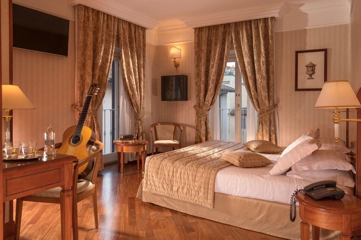 A hotel room with a bed, a desk with a guitar, a chair, a lamp, and a phone on the side table.