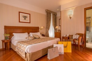 A hotel room with a large bed, a wooden headboard, a framed picture on the wall, a nightstand with a lamp, a chair, and a bathroom visible through an open door.