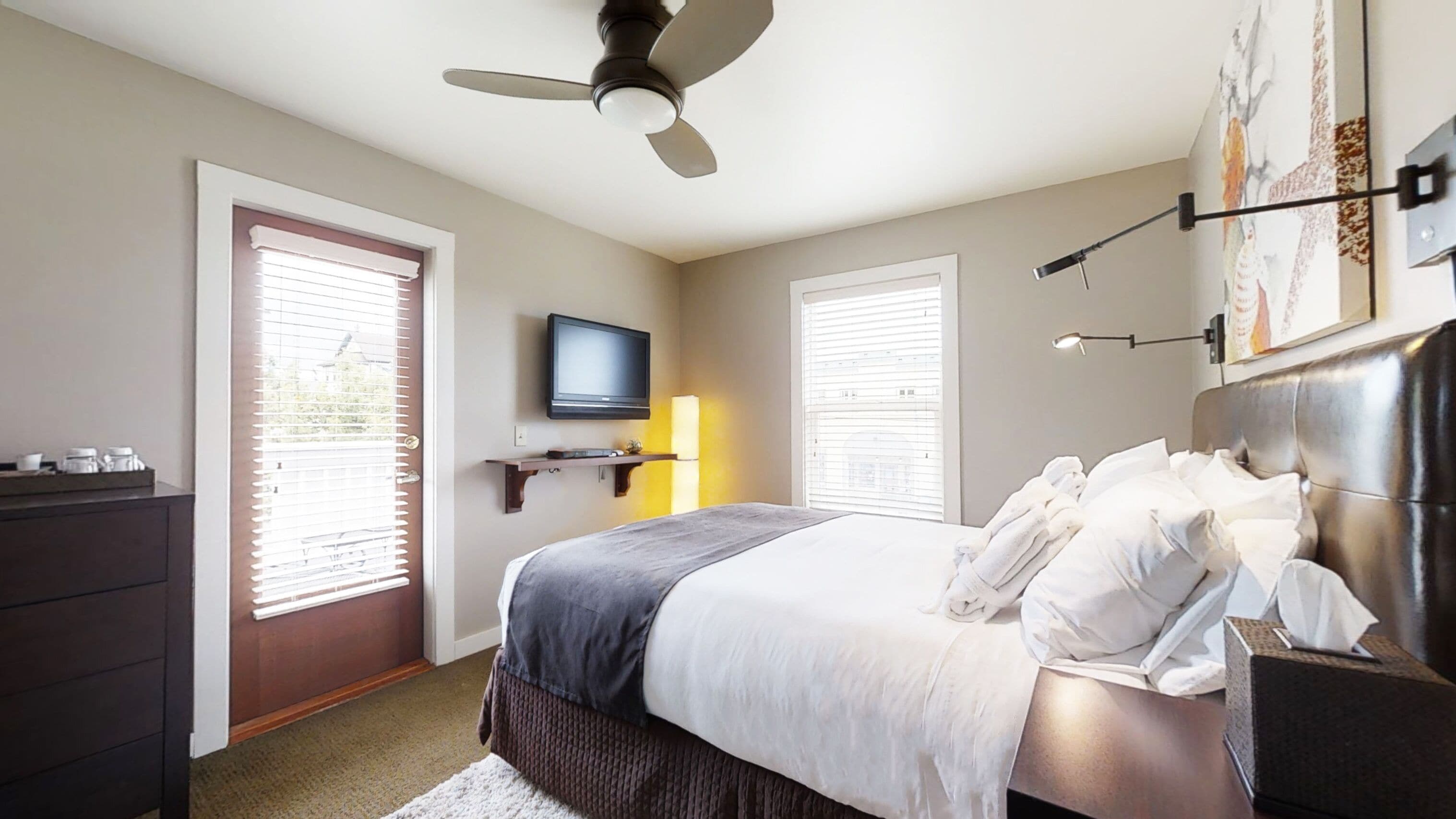 A modern bedroom with a bed, a ceiling fan, a TV, and a window with blinds.