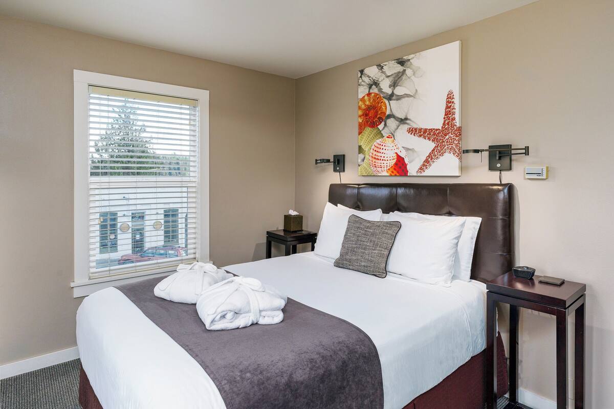 Una habitación de hotel con una cama grande, una ventana con persianas, una obra de arte colgada en la pared, una mesita de noche con lámpara y otro mueble auxiliar con teléfono.