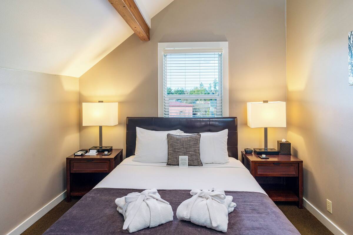 Habitación de hotel con una cama grande, dos mesitas de noche con lámparas, una ventana con persianas y dos toallas sobre la cama.