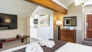 A hotel room with a bed, a TV, a painting, a wooden dresser, and a desk with a lamp. - Bird Rock Hotel (Friday Harbor)