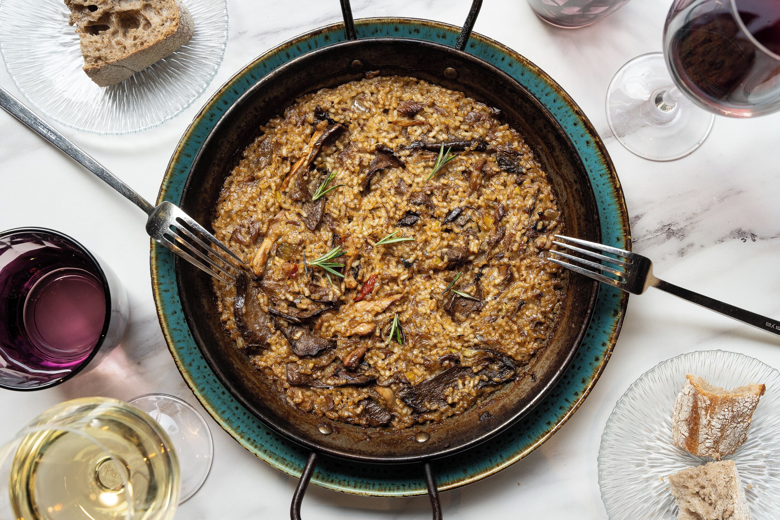 Una cazuela grande de risotto con champiñones y hierbas, acompañada de una copa de vino blanco y rebanadas de pan sobre una mesa de mármol.