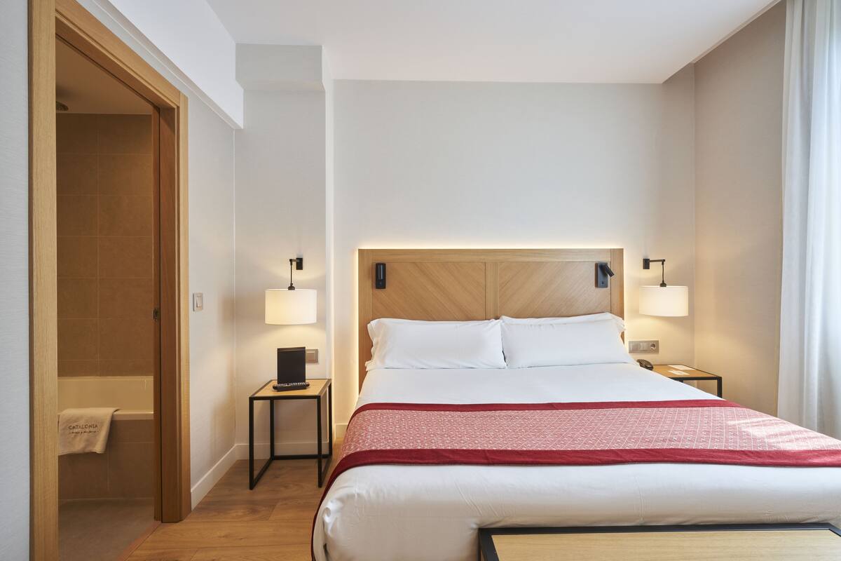 Quarto de hotel com cabeceira de madeira, cama grande com roupa de cama branca, duas luminárias de cabeceira, uma mesinha com abajur e porta de madeira que dá para um banheiro.