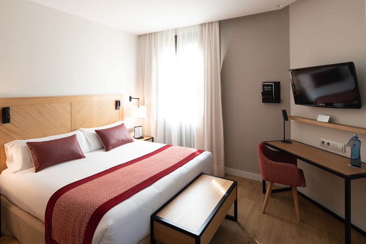 Quarto de hotel com uma cama, uma escrivaninha, uma cadeira, uma televisão e uma janela com cortinas.
