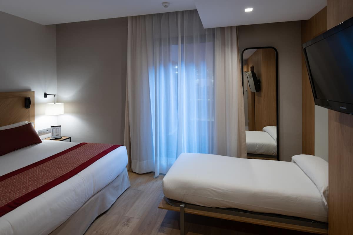 Quarto de hotel com uma cama grande, uma cama menor, mesa de cabeceira com luminária, televisão e janela com cortinas translúcidas.