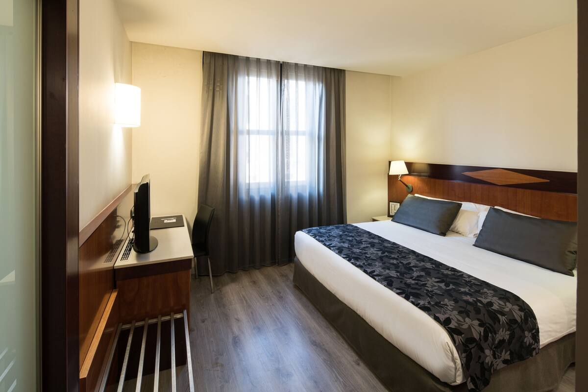 Quarto de hotel com cama, mesa de cabeceira, abajur e janela com cortinas.