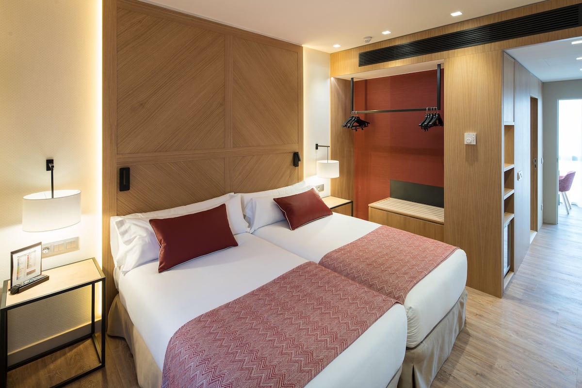 Quarto de hotel com duas camas, cabeceira de madeira, mesa de cabeceira com abajur e um espaço de estar com uma cadeira e uma mesa.
