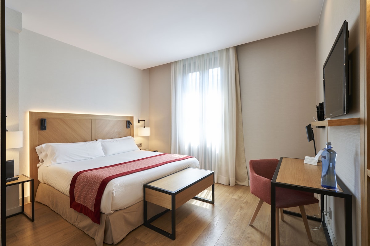 Quarto de hotel com cama, mesas de cabeceira, escrivaninha com cadeira, televisão e janela com cortinas.