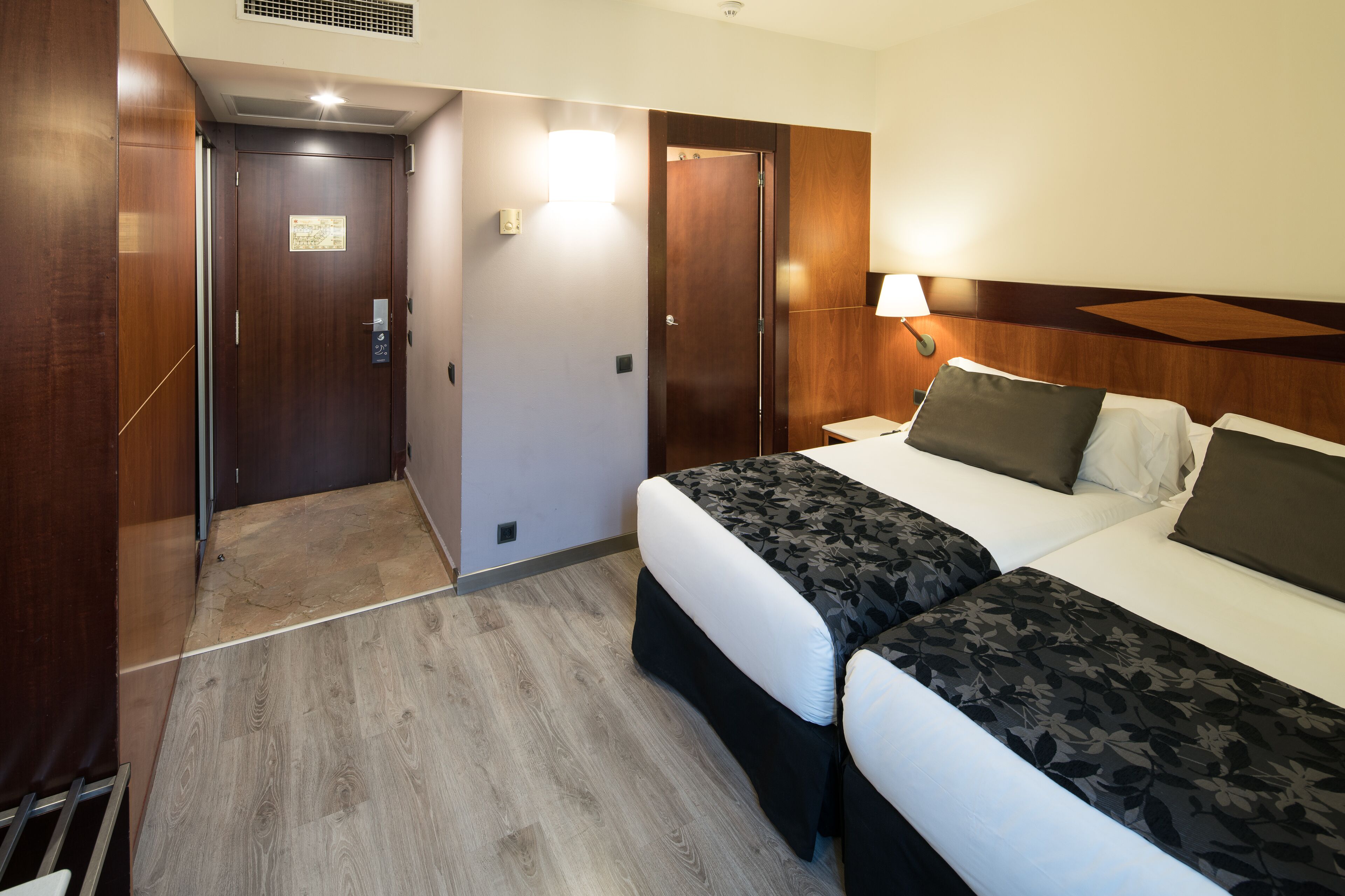 Une chambre d’hôtel avec une armoire en bois, un lit recouvert d’une couvre-lit à motifs floraux, une table de chevet avec une lampe et une petite fenêtre.