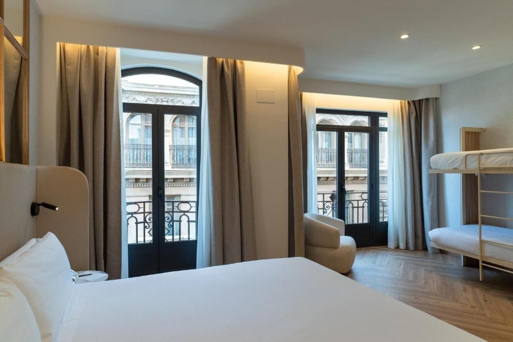 Habitación de hotel con una cama, una silla, literas y ventanas grandes con cortinas.