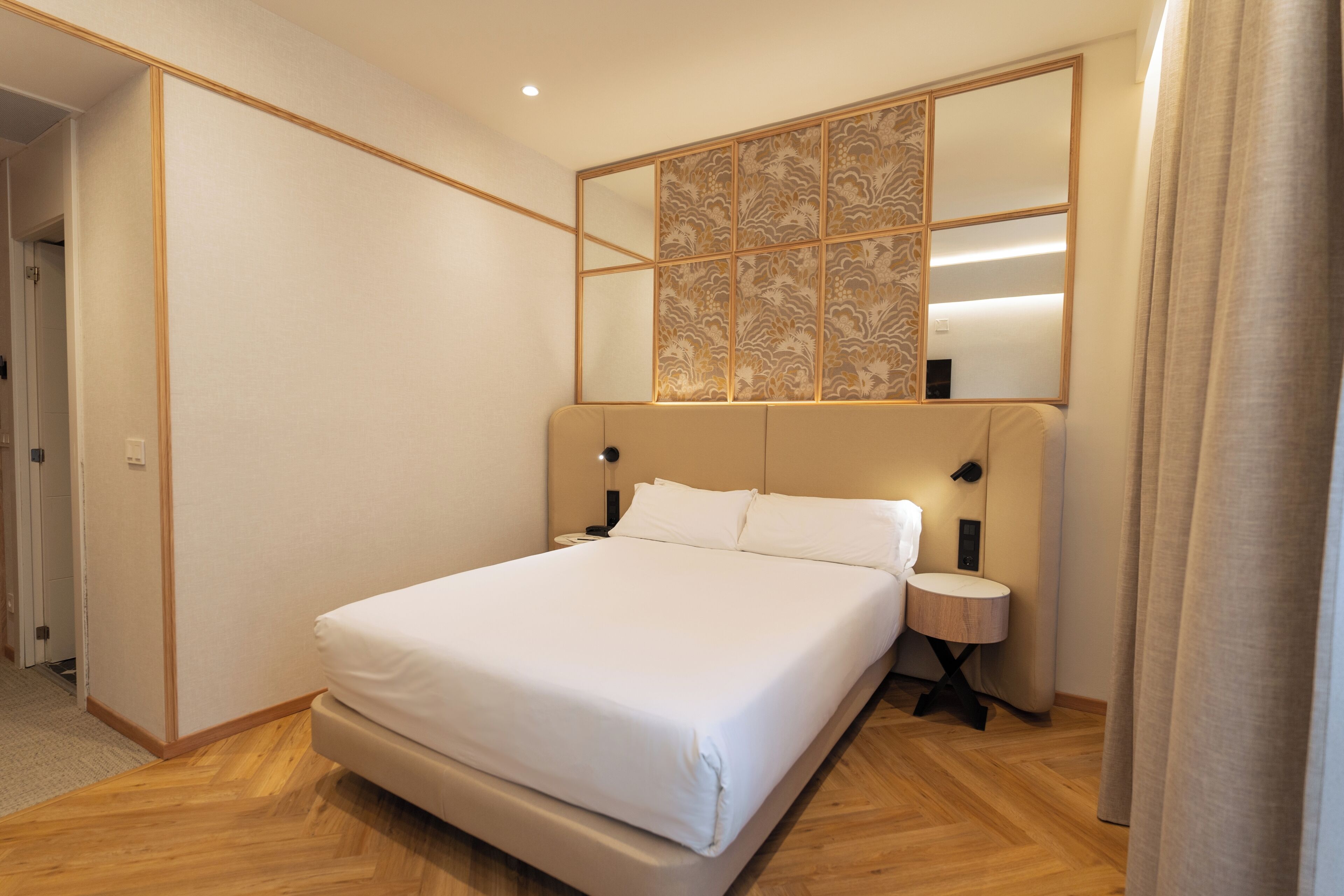 A modern hotel room with a large bed, a bedside table, a wall-mounted mirror, and a wooden floor.