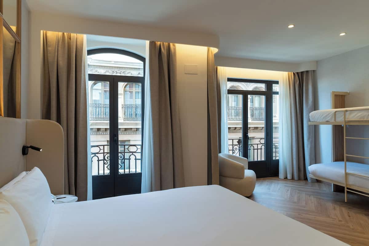 A hotel room with a large bed, a chair, a bunk bed, and a view of a balcony with wrought iron railings.